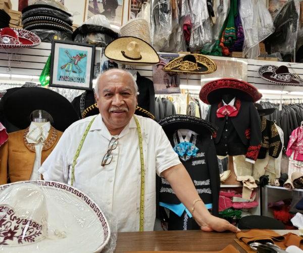一个面带微笑的留着小胡子的男人站在商店的柜台后面，戴着墨西哥流浪乐队的帽子.